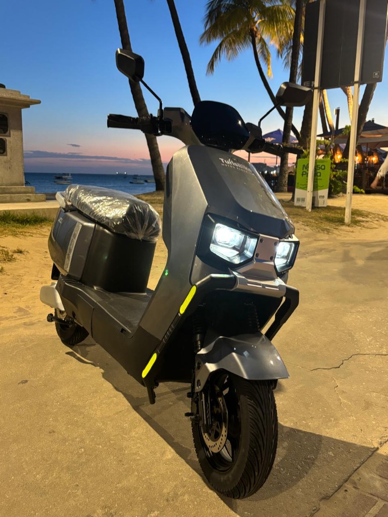 Moto elétrica cinza da TurisBike estacionada na orla ao entardecer em Maceió